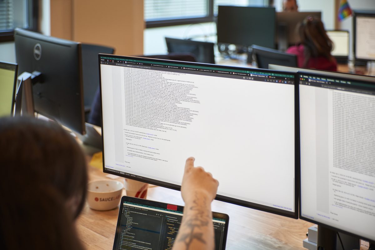 Eine Person zeigt auf einen Computermonitor mit Code, daneben ein Laptop und eine Tasse mit 'SAUCELABS' Logo.