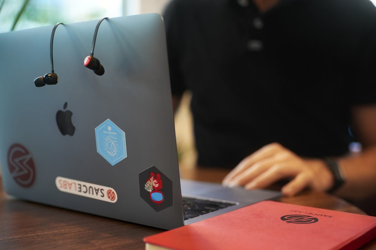 Ein Laptop mit Aufklebern und Kopfhörern, daneben ein rotes Notizbuch, im Hintergrund eine Person.