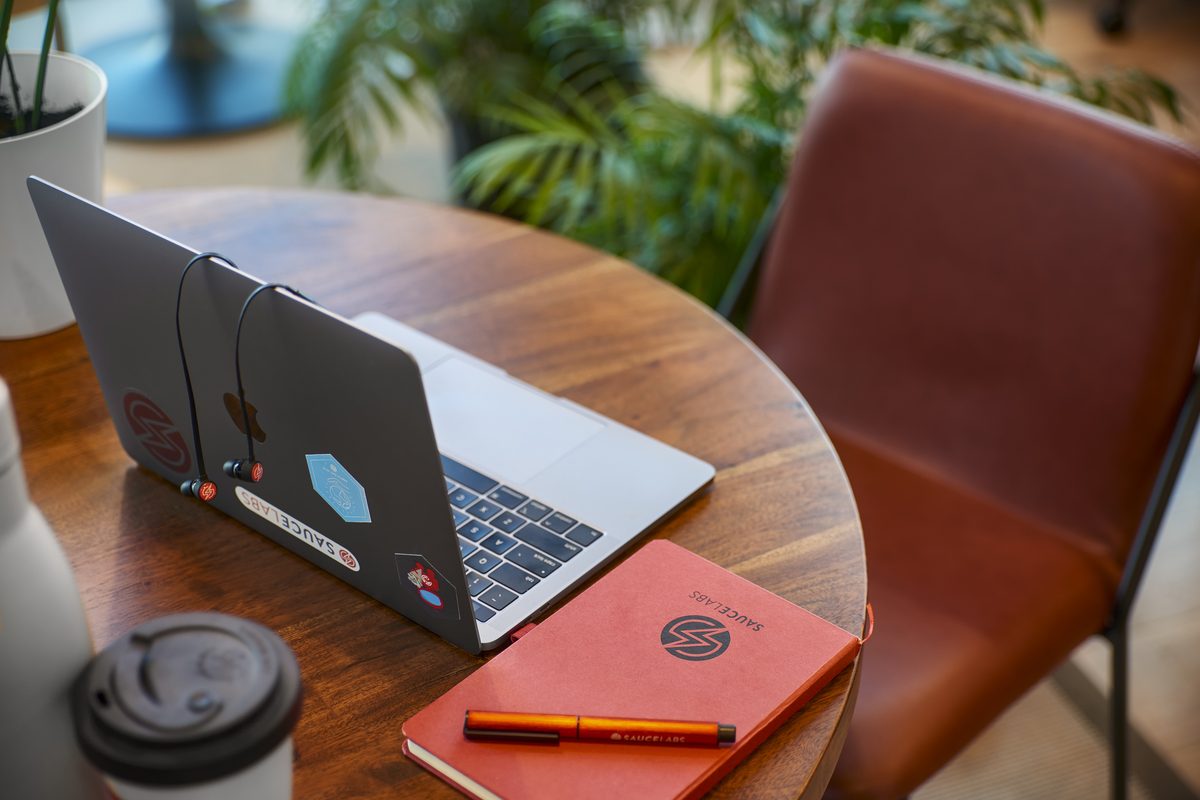 Ein Laptop mit Aufklebern, Kopfhörern, Notizbuch, Stift und Kaffeebecher auf einem Holztisch neben einem braunen Stuhl.