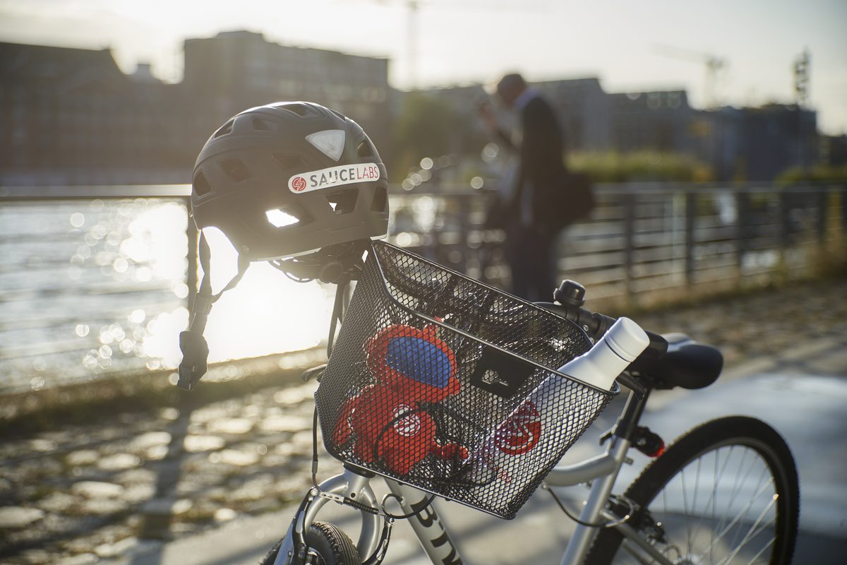 Ein schwarzer Fahrradhelm mit SauceLabs-Logo hängt an einem Fahrradlenker mit Korb, im Hintergrund ein Fluss und eine Person.