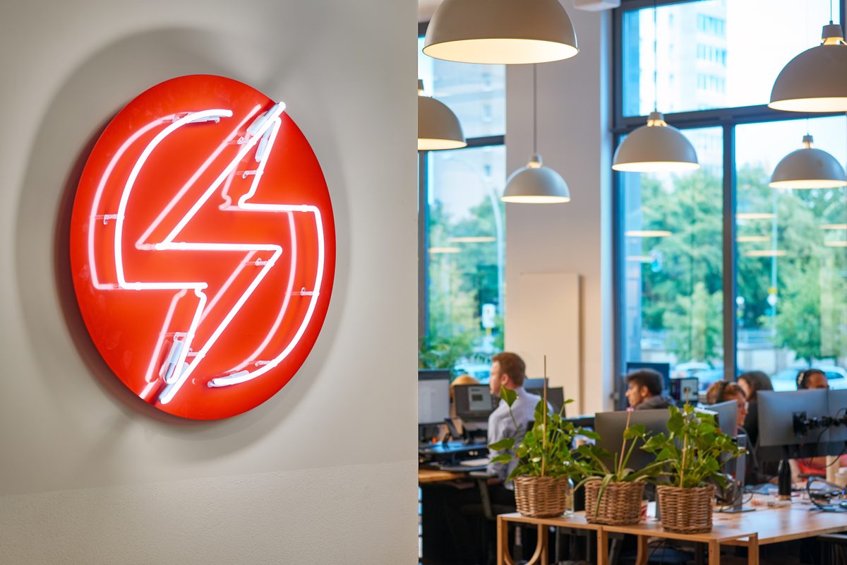 Rotes Neon-Logo an der Wand und ein offenes Büro mit Mitarbeitern, Pflanzen und Fenstern.
