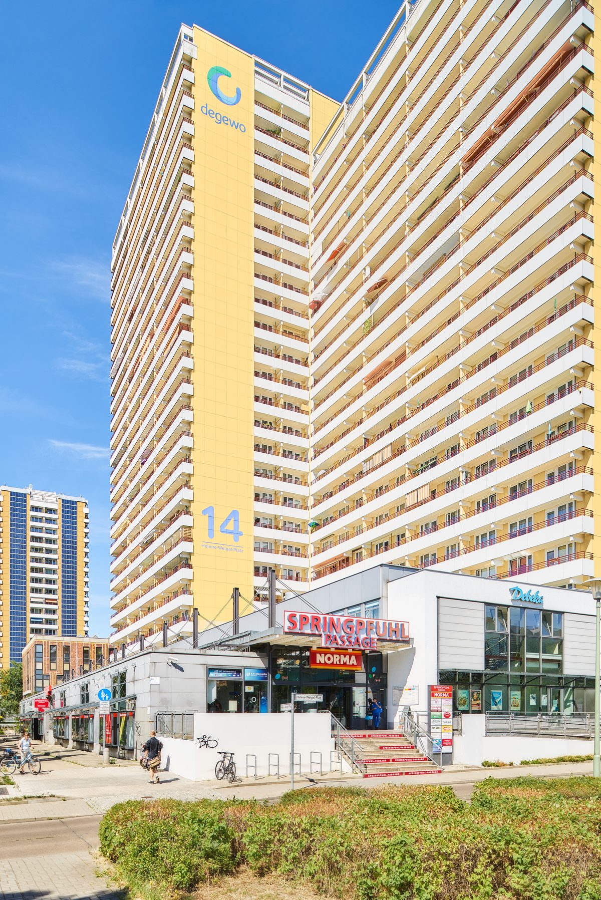 Gelbes Hochhaus mit vielen Balkonen und einer Ladenzeile im Erdgeschoss, davor eine Grünfläche und ein Gehweg.