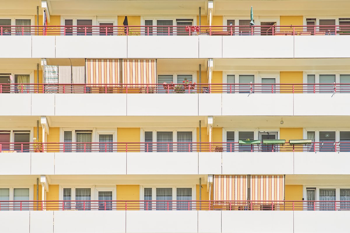 Fassade eines gelben Wohnhauses mit weißen Balkonen und roten Geländern, teilweise mit Markisen und Pflanzen.