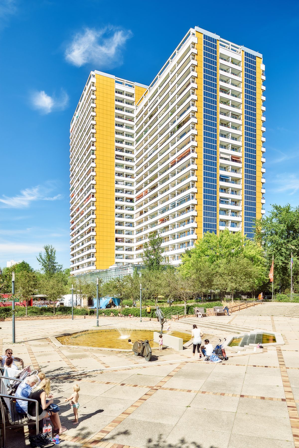Ein hohes Wohngebäude mit gelben und blauen Fassaden überblickt einen Platz mit Brunnen und Menschen.