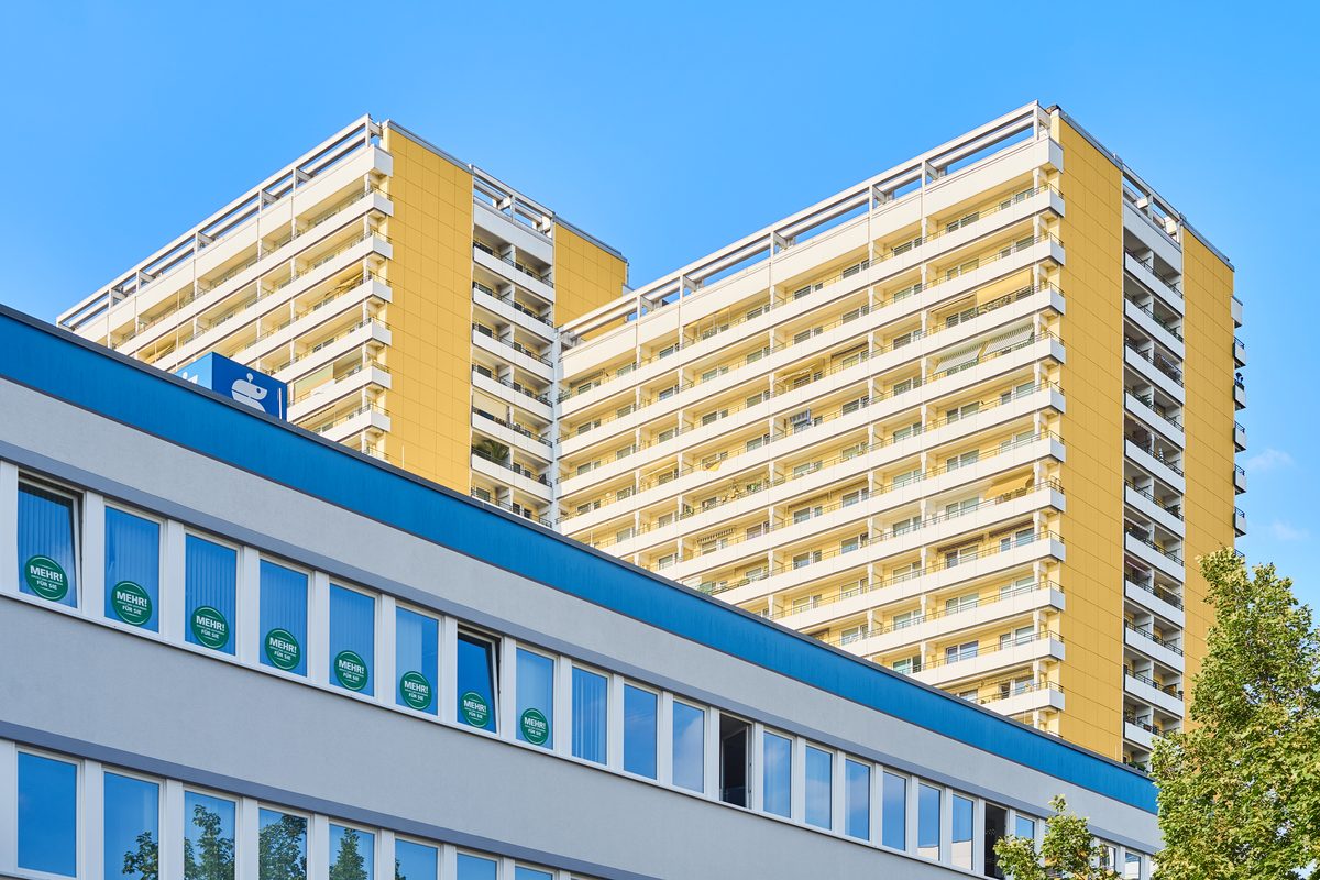 Zwei hohe Wohngebäude mit gelben Fassaden und Balkonen hinter einem niedrigeren Gebäude mit blauer und grauer Fassade.