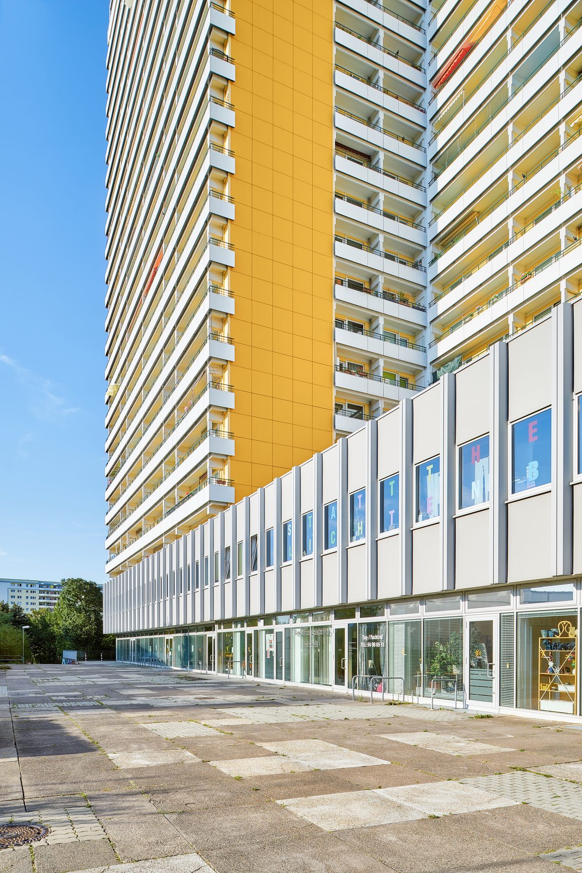 Ein hohes Wohngebäude mit gelber Fassade und vielen Balkonen über einem niedrigeren Gebäudeteil mit Geschäften.