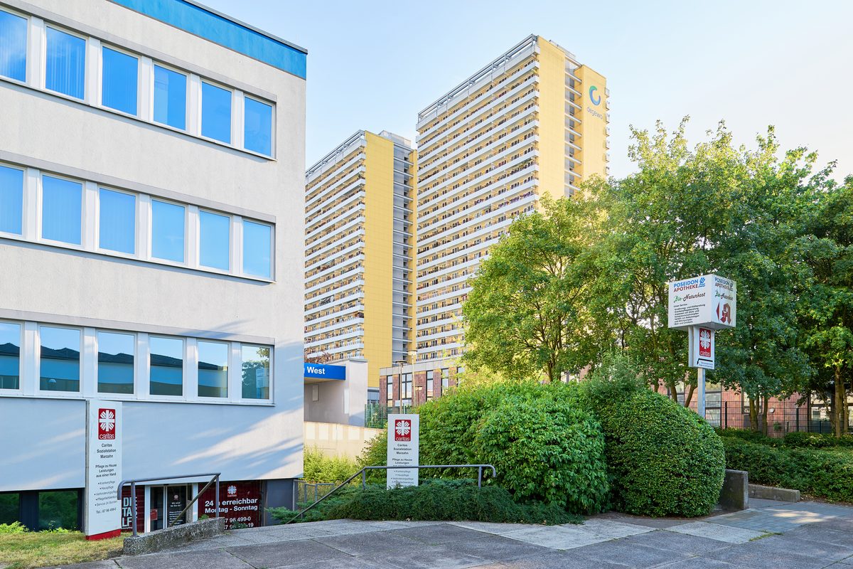 Zwei hohe gelbe Wohngebäude und ein weißes Gebäude mit blauen Akzenten stehen zwischen Bäumen und Büschen in Berlin-Marzahn.