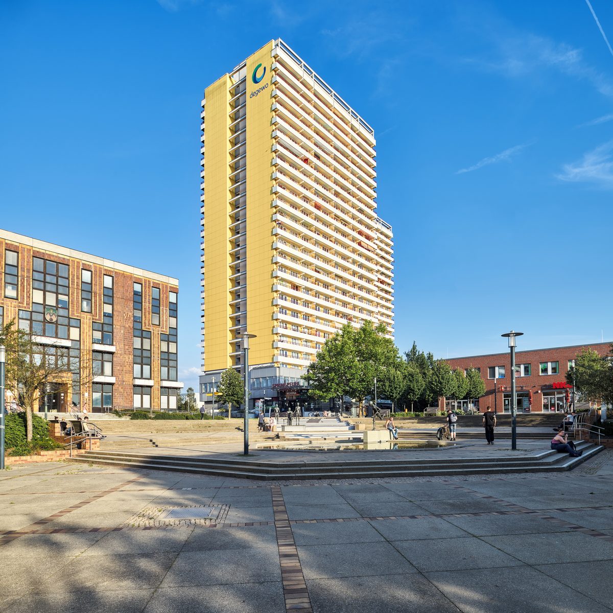 Ein gelbes Hochhaus mit Balkonen, umgeben von niedrigeren Gebäuden und einem Platz mit Menschen und Bäumen.