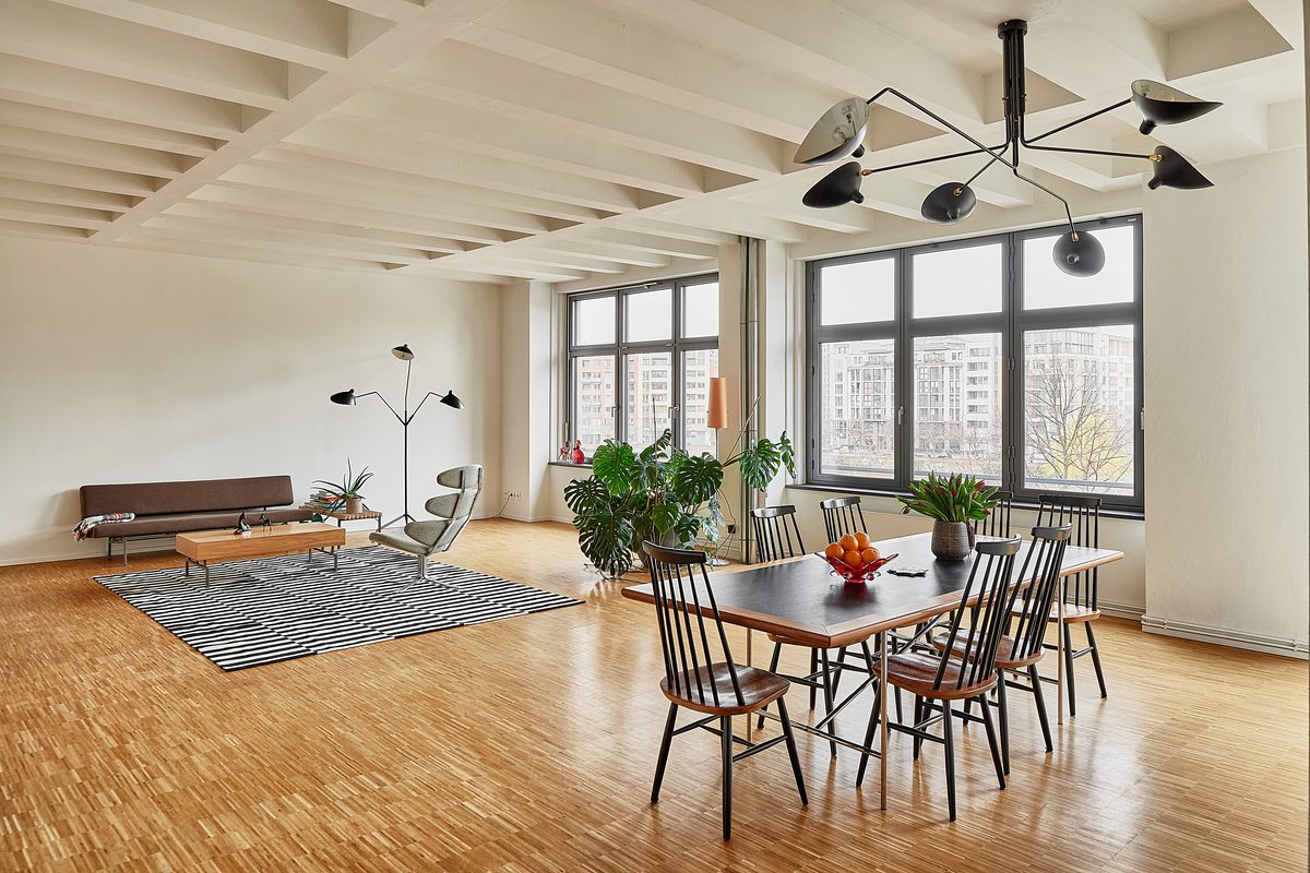 Ein großer Raum mit Holzboden, weißen Wänden, einem Esstisch, einem Sofa und großen Fenstern mit Blick auf Gebäude.