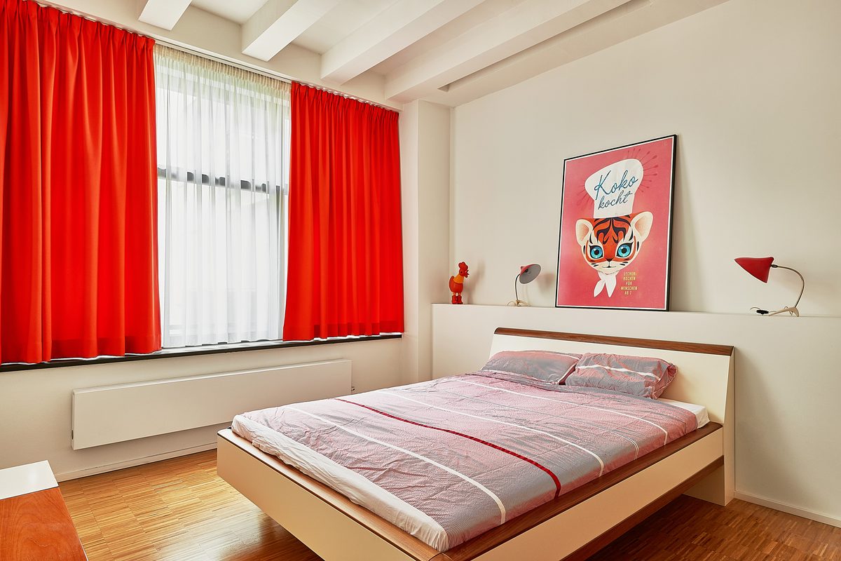 Ein Schlafzimmer mit Holzboden, einem Bett mit gestreifter Bettwäsche, roten Vorhängen und einem Poster an der Wand.